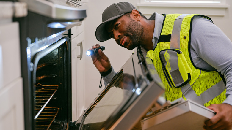 A repairman with a flashlight inspecting the inside of a kitchen oven.