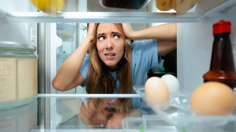 A woman biting her lip with her hands on her head as she stares into a fridge.