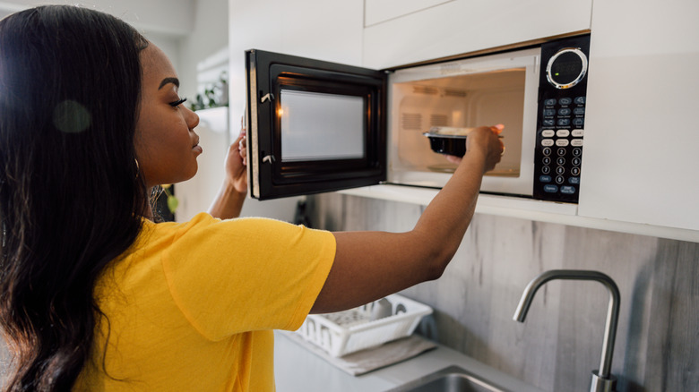 A woman putting a container of food in a wall-mounted microwave oven.