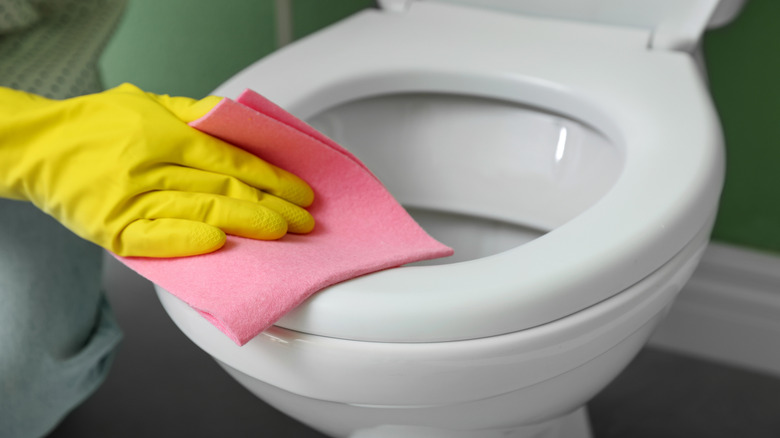 Person wearing yellow rubber gloves wipes down toilet seat with a pink cloth