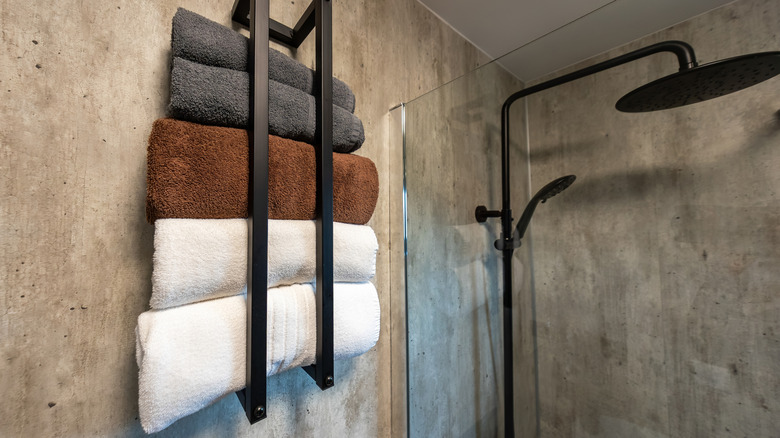 Two white, one brown, and two grey bath towels rolled up in a towel rack on a concrete bathroom wall near a glass shower with a black dual shower head.