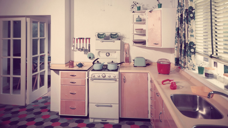 A 1950s kitchen is decked out with pink cabinets, a patterned floor, and floral curtains