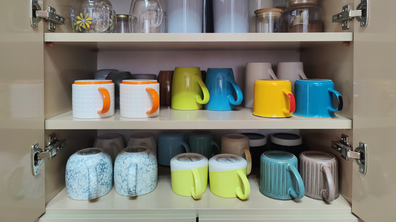 Two shelves in a kitchen cabinet filled with coffee mugs of all different shapes, sizes, and colors, most in pairs of two