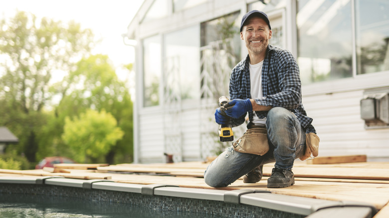 A man with a drill kneeling above the edge of a full pool, smiling