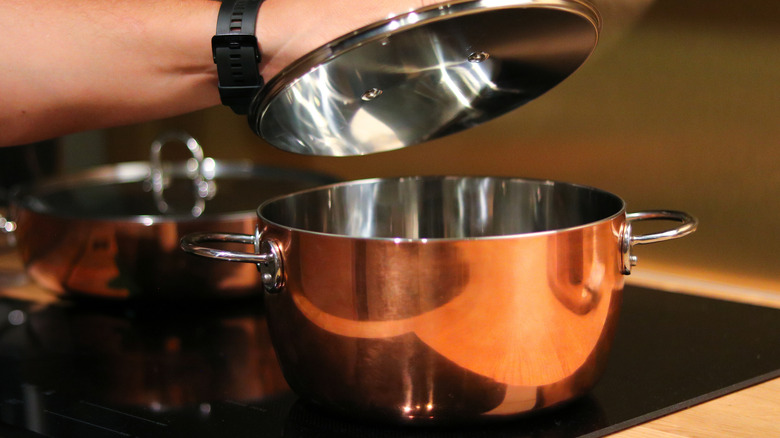 person lifting lid of shiny copper pot on glass cooktop