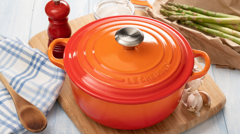 Red Le Creuset pot on a chopping board with garlic, asparagus and cooking items in the background