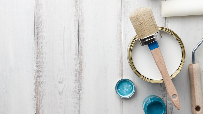 A can of white chalk paint on a wooden table with a brush on top, a roller next to it, and a small tin of aqua paint beside it