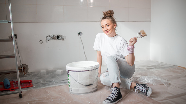 A person with a large bucket of paint and a paint brush in hand on a tiled floor in a bathroom