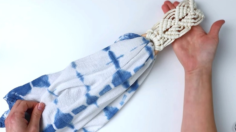 A person holds a tea towel holder made in a macramé technique and a tie dye white and blue tea towel.