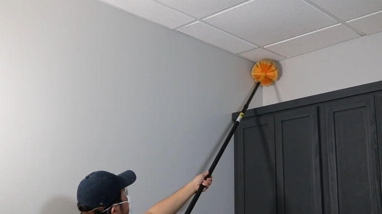 Man wearing a mask and cleaning away ceiling cobweb with orange cobweb duster