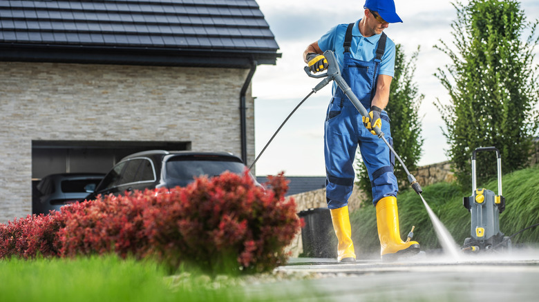 Man cleaning concrete with a pressure washer in blue overalls and hat, yellow rubber boots, and a