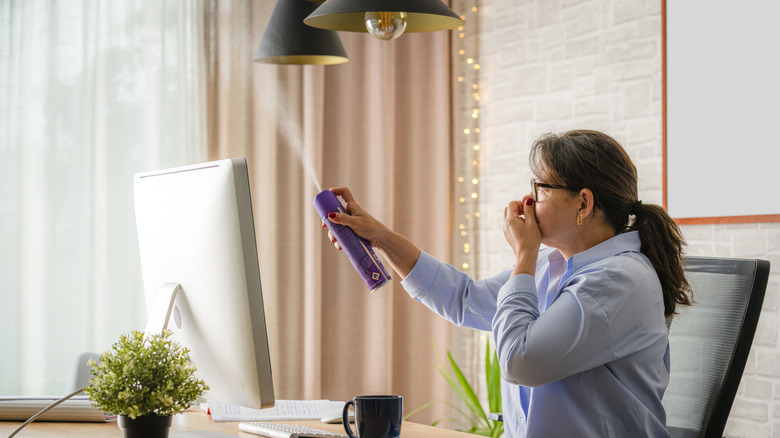 A woman at her desk is holding her nose while spraying room spray