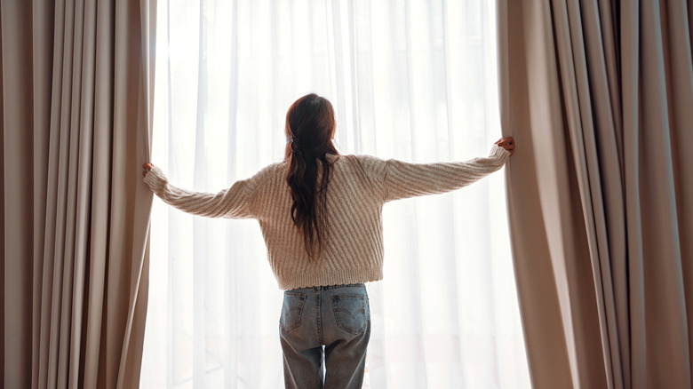 Woman pulls blackout curtains over sheer panels to block bright sunlight.