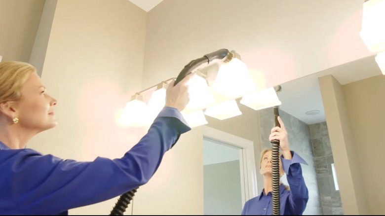 A woman uses a vacuum wand to clean a light fixture.
