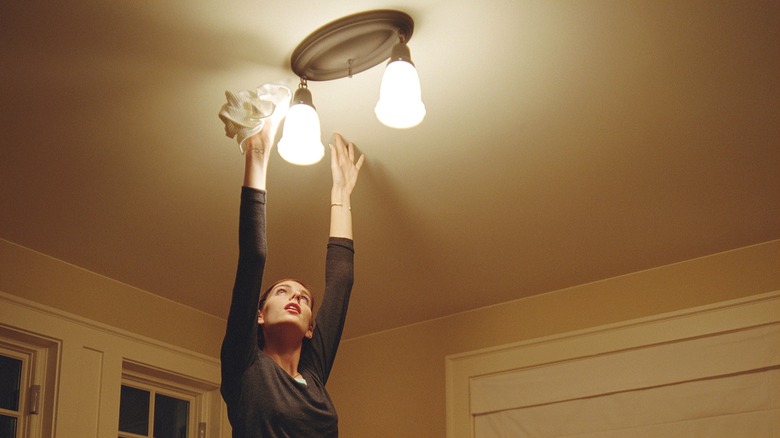 A woman struggles to reach and clean a light fixture.