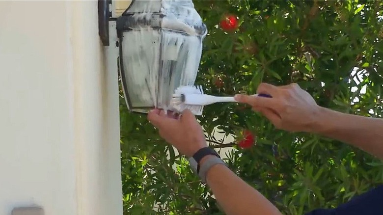 A man scrubs an outdoor light fixture with a household scrubbing brush.