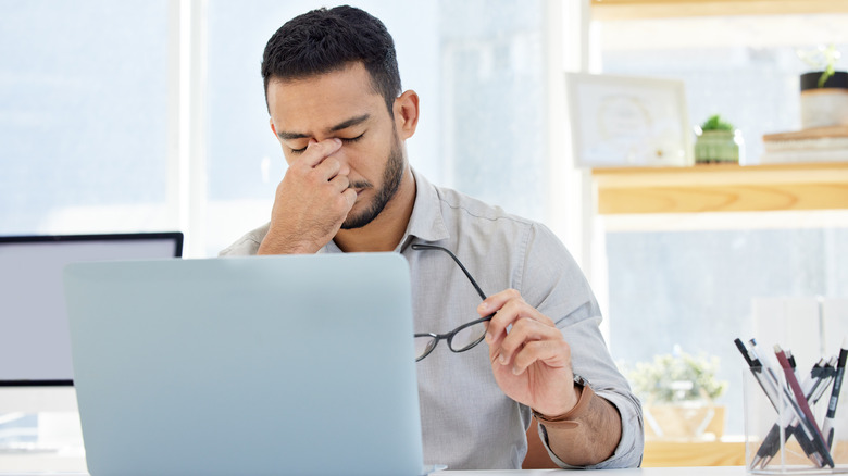 A man with a headache holds his glasses in one hand and pinches the bridge of his nose with the other.