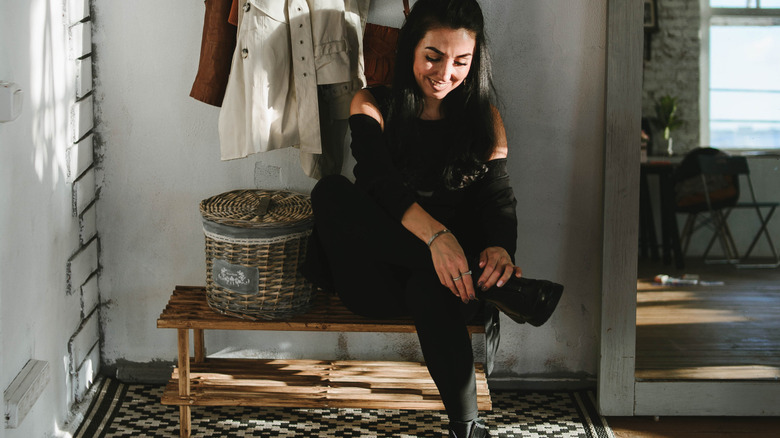 A woman sits on a bench to take her shoes off in the entryway to a home.