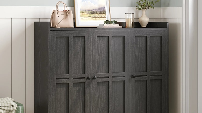 A dark shoe cabinet with items displayed on the tabletop sits against light-hued wainscoting in a home.