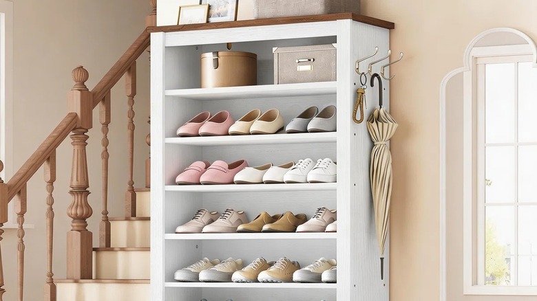 A slim, tall, open cabinet holding lots of different styles of shoes near a staircase and window.