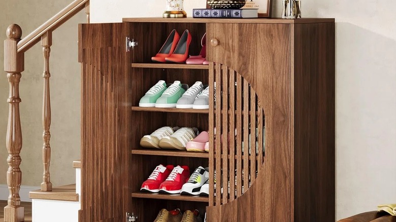 A brown wood-look shoe cabinet with a circular slats on the doors in a home hallway beside a staircase.