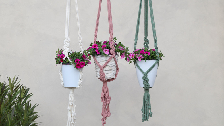 White, pink, and green crochet plant hangers in a room with a textured cream-gray wall and another houseplant.