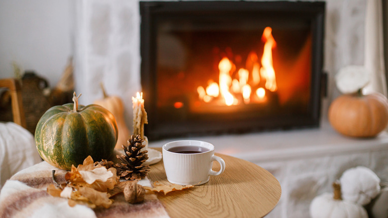 A cozy fall interior with a pumpkin, pinecone, and mug of coffee on a table, and a lit fireplace in the background.