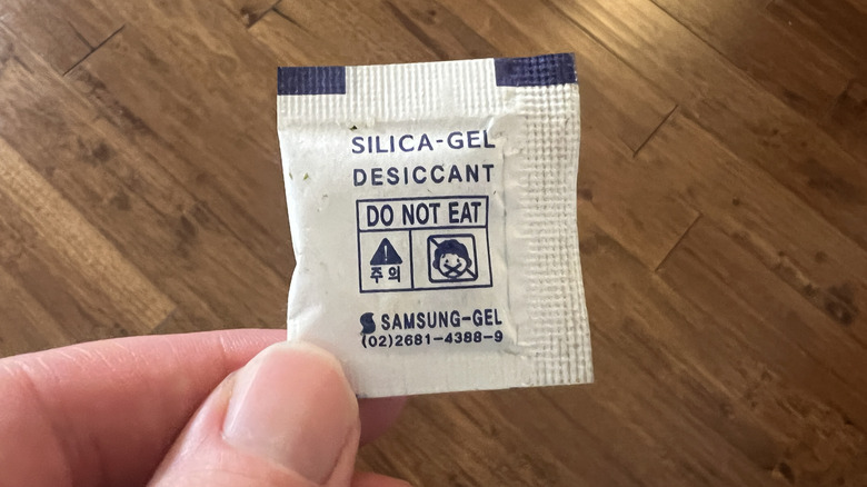 A person holds a silica gel packet in their hand above a wooden floor.