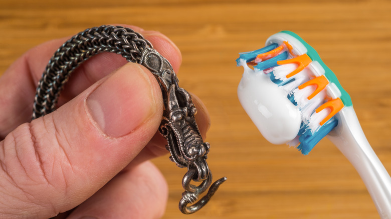 A person holds a toothbrush with toothpaste in one hand and a tarnished piece of silver jewelry in the other hand.