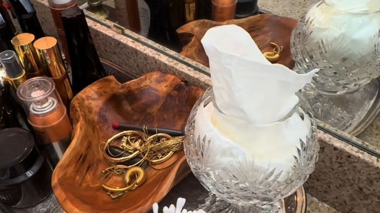 Tissues inside of a glass vase with embellishments, it sits on the bathroom counter next to a wooden bowl collecting jewelry