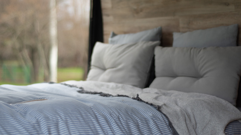A blue striped comforter on a bed with soft focus pillows and a wooded area outside the window behind the bed.