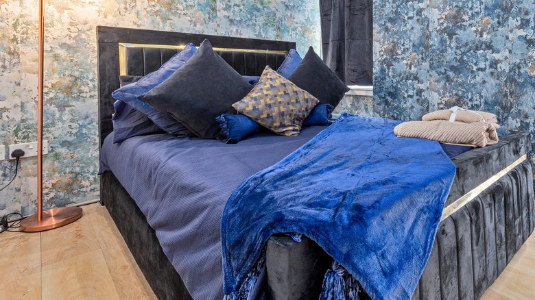 A navy velvet bed with various shades of blue bedding and a blue velvet comforter folded over the end of the bed. The walls are covered in an abstract blue smudged wallpaper.