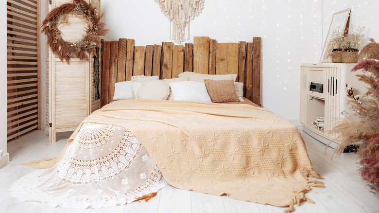 A bed covered in a vintage cream throw with a lace underlay. The bed has a driftwood headboard and there is a dried wreath on the wall.