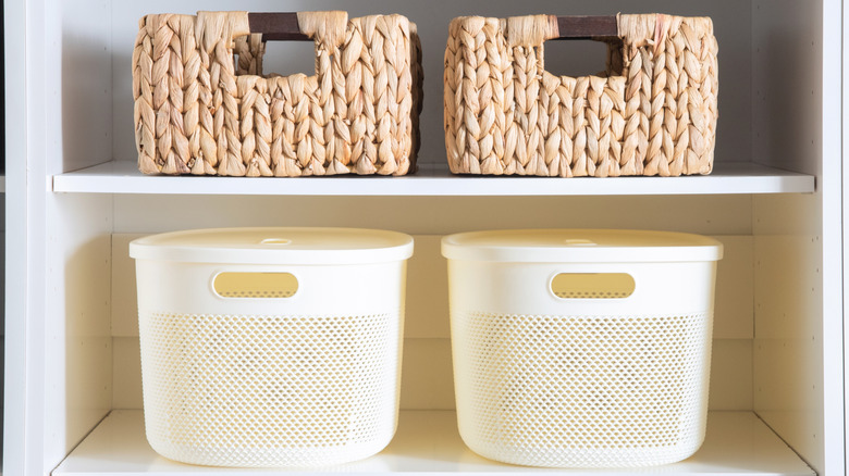 Scandinavian inspired closet shelf detail with woven baskets and cream plastic containers on white shelving, near gray patterned boxes and neutral clothing in a clean minimalist wardrobe setup.