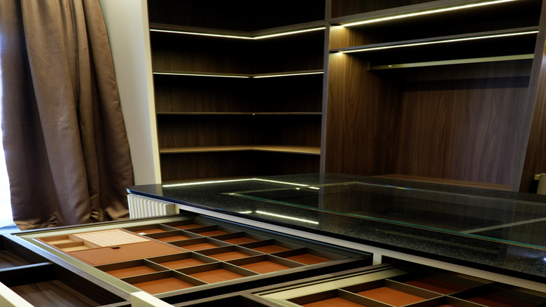 Luxury walk-in closet featuring a central island with a black granite top and glass display. Open drawers show organized jewelry compartments lined with brown leather and walnut wood finish.