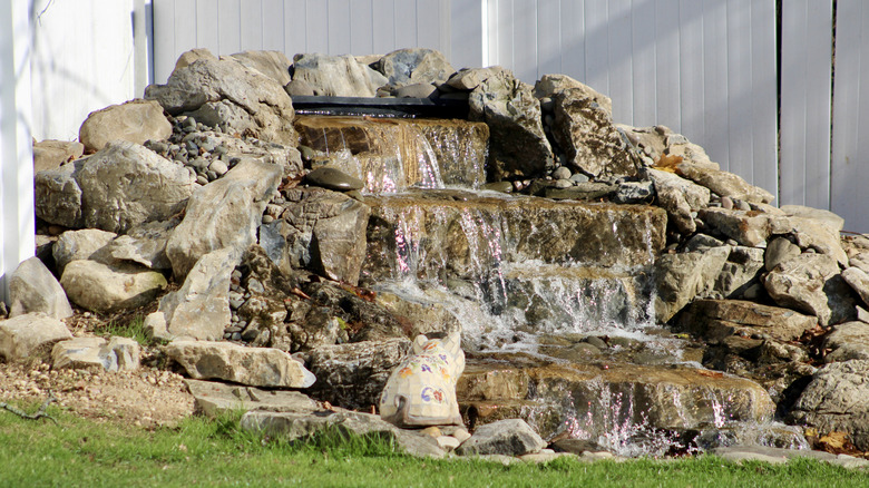 A small backyard pond with waterfalls over stones near a fence