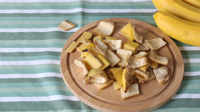 Chopped banana peels sit on top of a circular wooden plate with a bunch of bananas in the corner of the picture