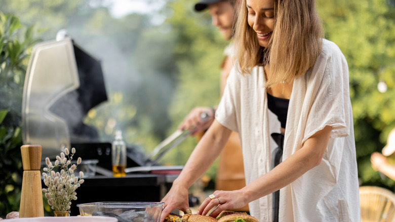 A woman getting food together while a man grills in background
