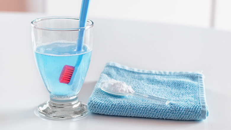 Denture cleaner in a cup with a toothbrush next to a washcloth and a spoon with powder on it