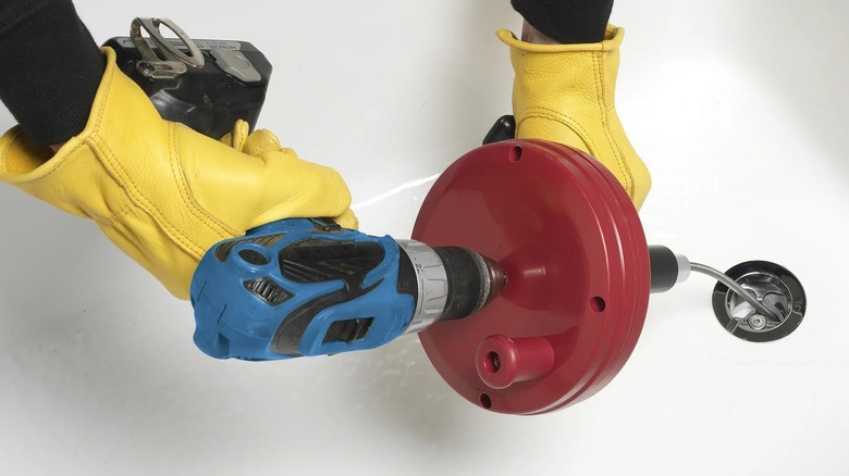 Hands with yellow gloves use an electric drill to turn a red hand-crank plumber's auger inserted into a drain.