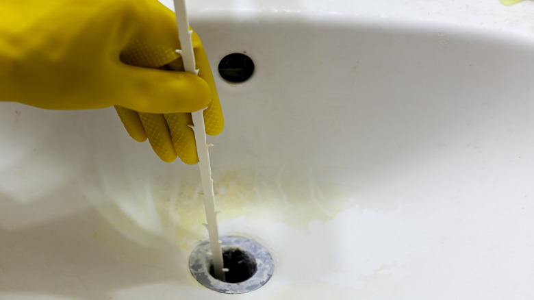 A white drain snake being pushed down a drain hole by a yellow-gloved hand.