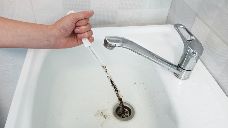 A hand pulls a dirty sink drain brush out of a bathroom sink drain.