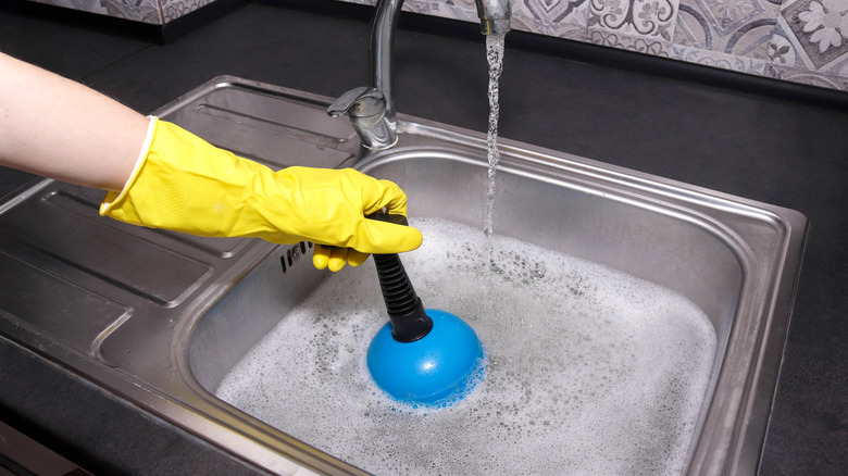 A yellow-gloved hands used a blue plunger to unclog a sink.