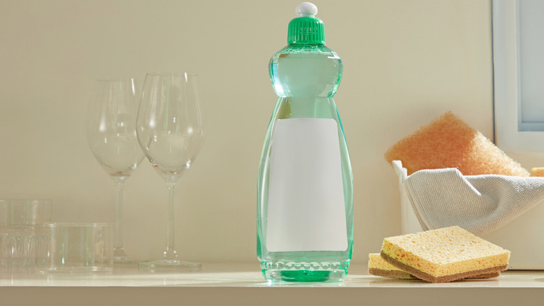 A bottle of dish soap on a shelf with wine glasses and sponges