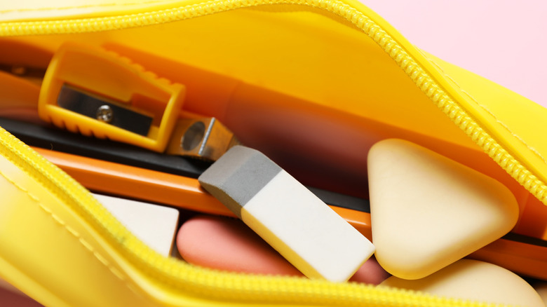 An eraser sitting in a bright yellow pencil case with sharpeners, pencils, and pens