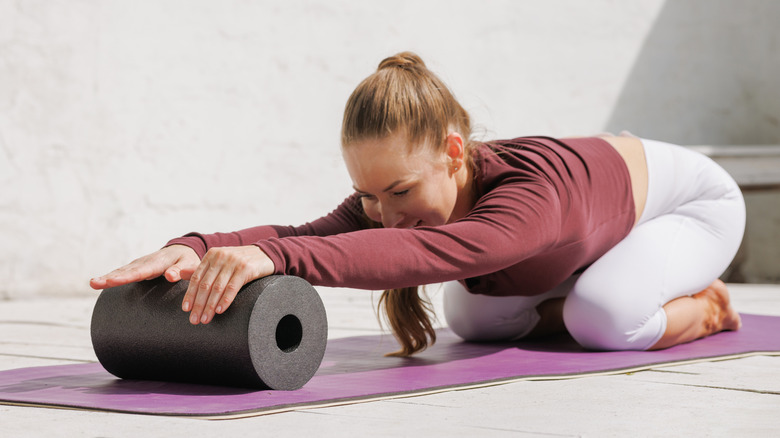A woman with white pants, a maroon long sleeved shirt, and a high ponytail using a roller ball on a purple yoga mat