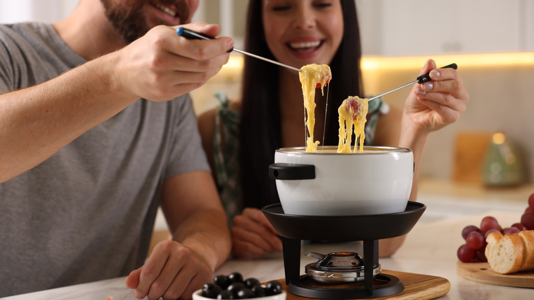 A fondue pot filled with cheese being used by a couple on a countertop with trays of food around it