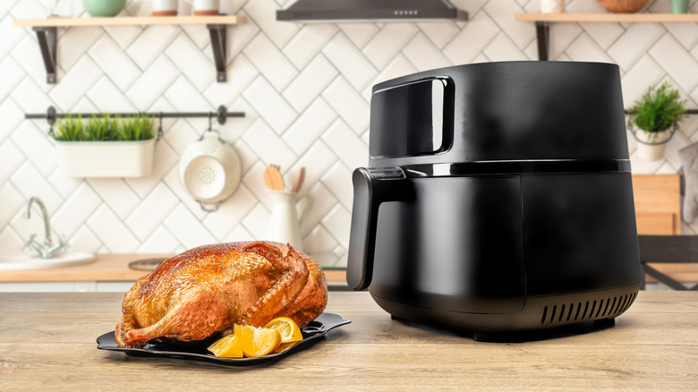 A black air fryer on a wooden counter with a golden-brown whole fried chicken beside it.