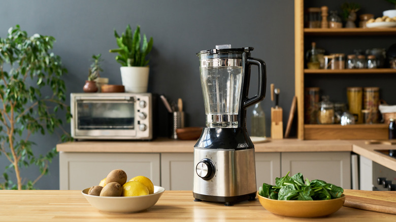 A wooden kitchen counter with a blender and bowls of fruit and vegetables on it. A toaster oven is in the background.