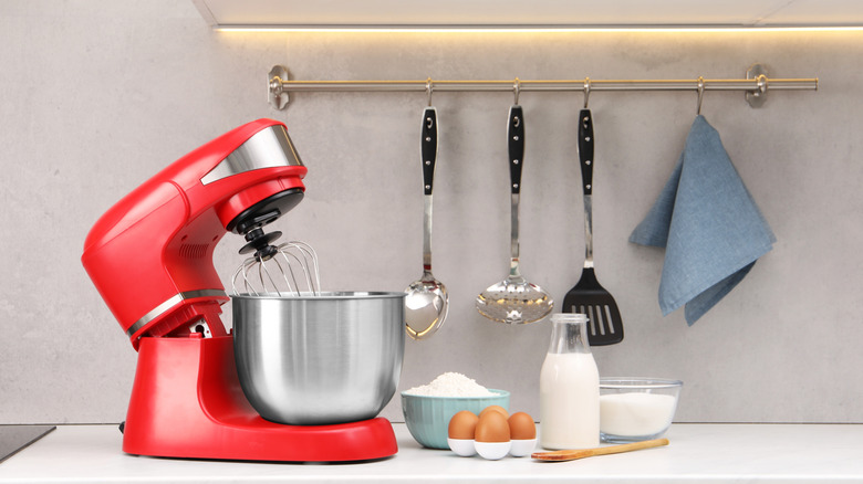 A red stand mixer on a kitchen counter with baking goods like milk, flour and eggs beside it, and utensils hanging on a rack on the backsplash.